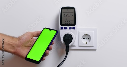Man switches on wattmeter and controls electrical device power consumption and examines functioning looking at smartphone. Person tries to fix problem photo
