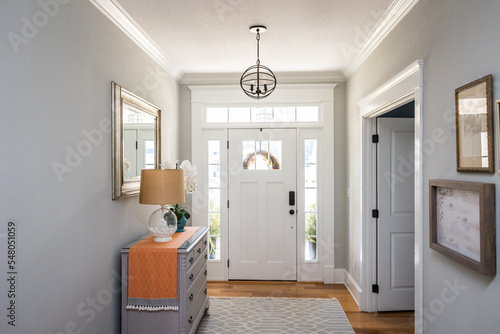 An open large and wide interior front door hallway foyer with transom, hanging light fixture, coastal colors and entry way table and wood floors photo