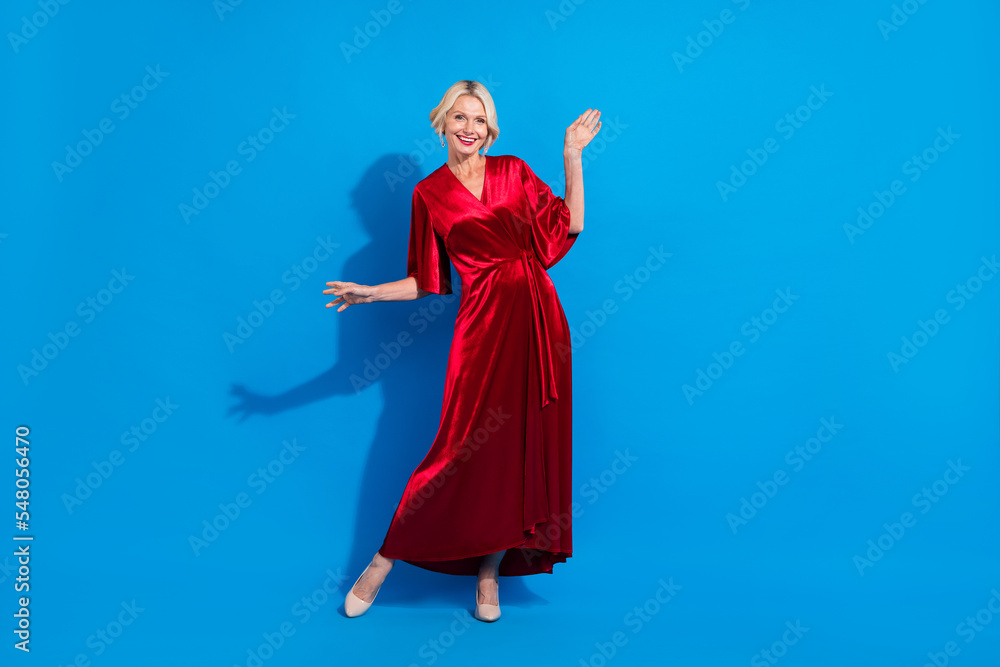 Full length photo of friendly sweet senior lady wear red shiny dress waving hand having fun isolated blue color background Full length photo of friendly sweet senior lady wear red shiny dress waving hand having fun isolated blue color background