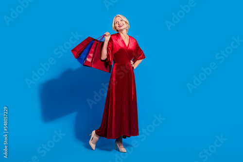 Full length photo of pretty dreamy lady dressed shiny red clothes holding shoppers looking empty space isolated blue color background
