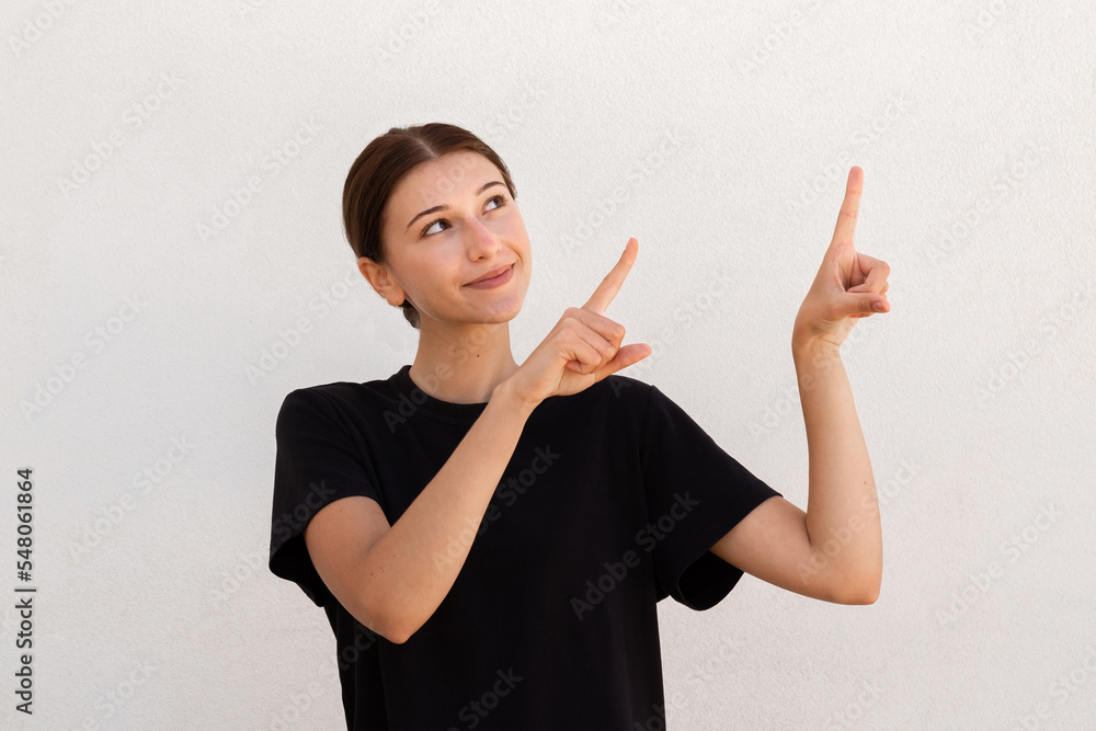 Portrait of positive young woman pointing forefingers upwards over white background. Caucasian woman wearing black T-shirt smiling advertising something. Promotion concept Portrait of positive young woman pointing forefingers upwards over white background. Caucasian woman wearing black T-shirt smiling advertising something. Promotion concept