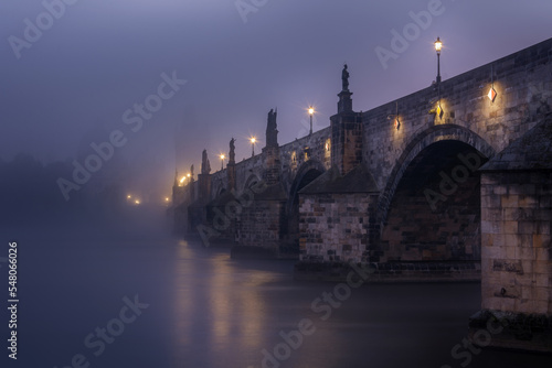 Charles bridge from below in the fog in the early morning in Prague with statues and lanterns on the bridge. Czech Republic.