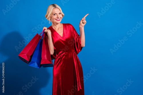 Photo of sweet adorable senior woman wear red shiny dress holding shoppers pointing empty space isolated blue color background