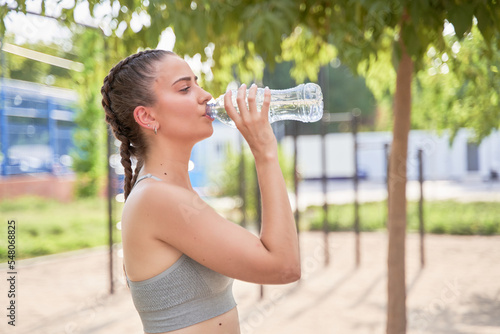 young Caucasian athlete with braids drinking water after outdoor training.
