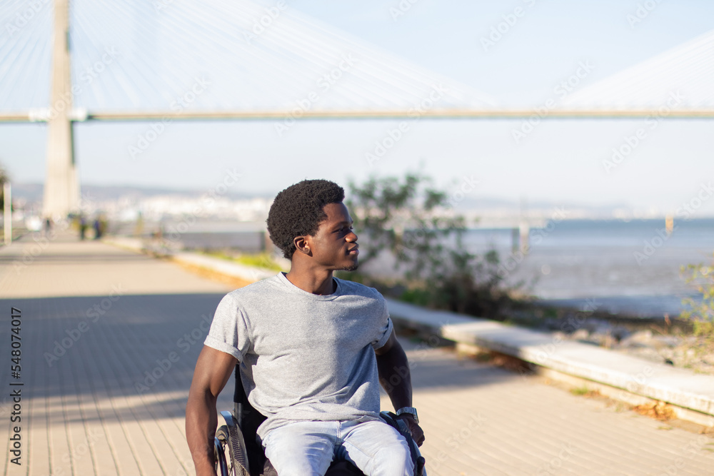 Fototapeta premium Happy Black man in wheelchair enjoying sea view while riding along park road near seashore. Smiling African American guy with disability spending time outside. Disability, lifestyle concept.