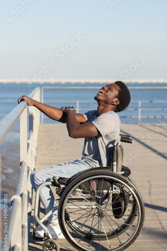 Cheerful Black man sitting in wheelchair at seafront on sunny day. Side view of confident guy with disability holding railing with his hand, and laughing happily. Disability, attitude concept.