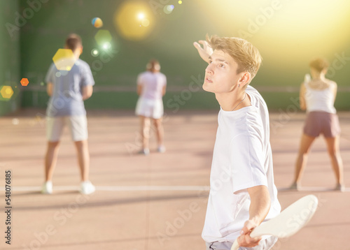 Focused young man playing friendly pelota goma match on outdoor summer court with fronton wall, swinging wooden paleta to hit small rubber ball © JackF