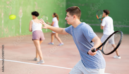 Sporty young man playing popular team game frontenis at open-air fronton court on summer day, ready to hit rubber ball with racquet