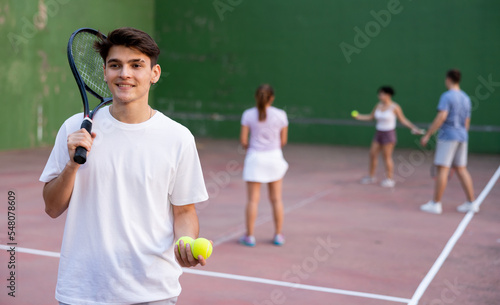 Portrait of confident positive young hispanic frontenis player standing on summer day on open-air fronton court after friendly match, holding racket and ordinary yellow tennis balls