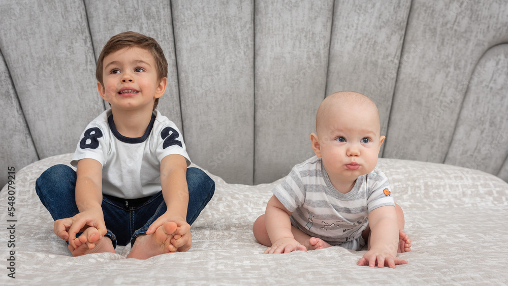 Close-up portrait, two brothers sitting on the bed and looking away, smiling, playing. The view is straight. Two little boys sitting on a bed. Baby boy and his younger brother together on the bed Close-up portrait, two brothers sitting on the bed and looking away, smiling, playing. The view is straight. Two little boys sitting on a bed. Baby boy and his younger brother together on the bed