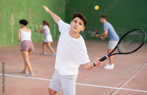 Athletic young hispanic guy playing frontenis on open court on summer day, hitting ball with strung tennis racquet to score to opposing team. Popular sports
