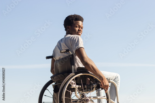 Black man in wheelchair enjoying sunny day in park, looking back and smiling at camera over the shoulder. Young African American guy with disability having fun outdoors. Disability, motivation concept