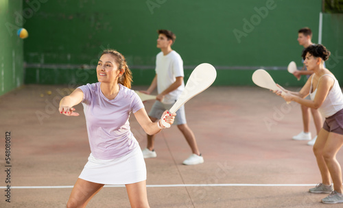Hispanic woman pelota player hitting ball with wooden racket during training game on outdoor Basque pelota fronton. © JackF