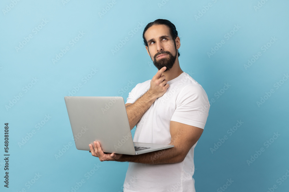 Portrait of man with beard wearing white T-shirt holding laptop and rubbing chin looking away, thinking over new business project. Indoor studio shot isolated on blue background. Portrait of man with beard wearing white T-shirt holding laptop and rubbing chin looking away, thinking over new business project. Indoor studio shot isolated on blue background.