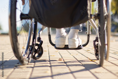Person of unrecognizable gender and age in wheelchair spending time outside. Closeup back view shot of feet in white sneakers and wheels. Disability concept. photo
