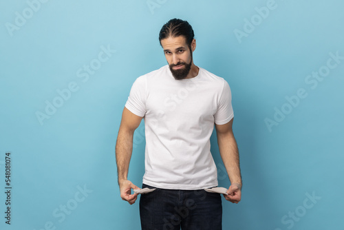 Portrait of man with beard wearing white T-shirt showing empty pockets and looking frustrated about loans and debts, has no money, jobless. Indoor studio shot isolated on blue background. photo