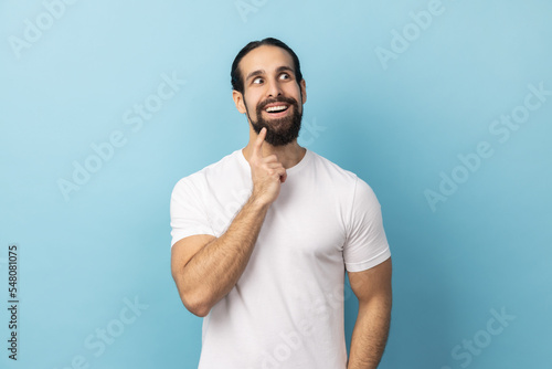Portrait of man with beard wearing white T-shirt looking away with pensive expression, dreaming pleasant thoughts, fantasizing and making wish. Indoor studio shot isolated on blue background.