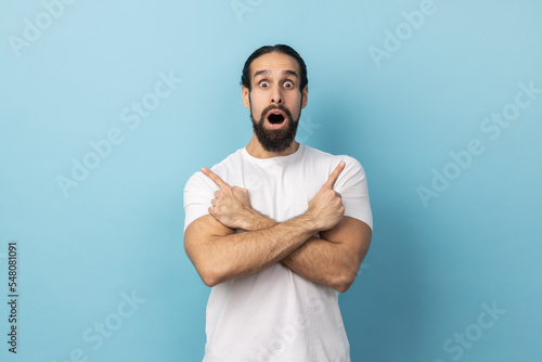 Portrait of amazed surprised man with beard wearing white T-shirt standing with crossed hands and pointing to copy space on both sides. Indoor studio shot isolated on blue background.