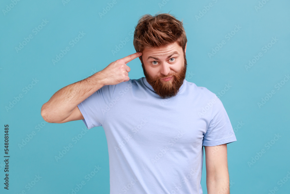 Portrait of young bearded man showing stupid gesture with finger near head, out of mind, accusing crazy dumb plan, reckless expression. Indoor studio shot isolated on blue background. Portrait of young bearded man showing stupid gesture with finger near head, out of mind, accusing crazy dumb plan, reckless expression. Indoor studio shot isolated on blue background.