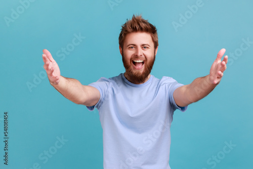 Free hugs, come into my arms. Portrait of happy bearded man stretching hands to camera and smiling broadly, going to embrace, share love. Indoor studio shot isolated on blue background.