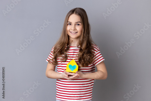 Portrait of little girl wearing striped T-shirt holding in hands little paper house looking at camera with smile, dreaming about apartment. Indoor studio shot isolated on gray background. © khosrork