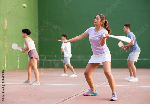 Positive fit young Latina playing pelota on traditional open court with front wall on summer day, hitting ball with wooden racket to score to opposing team © JackF
