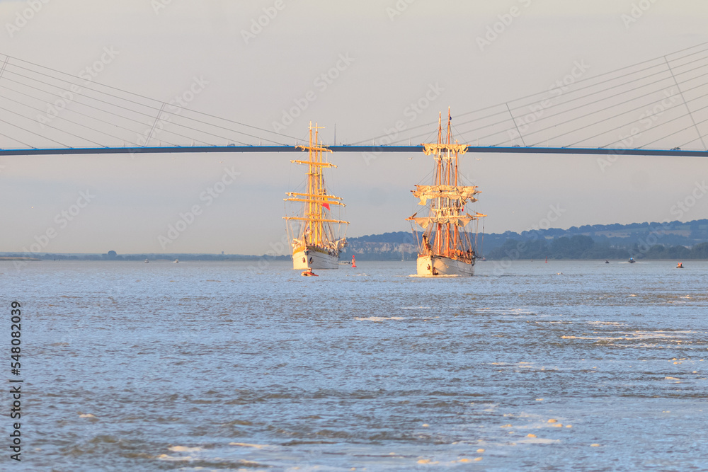 HONFLEUR, NORMANDY, FRANCE: Armada 2019 Grande Parade, tall ships Mircea and Le Français sailing in the Seine Estuary, in front of Normandy Bridge, with warm evening light - obrazy, fototapety, plakaty 