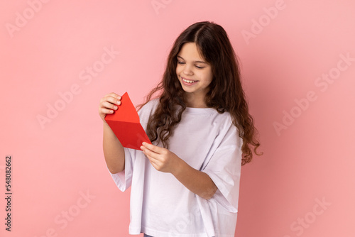 Billede på lærred Portrait of little girl wearing white T-shirt reading letter or greeting card, holding envelope, smiling and rejoicing pleasant news