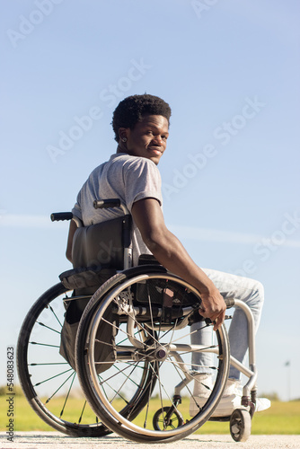 Black man in wheelchair enjoying warm day in park, looking back at camera and smiling. Short-haired African American guy with disability having good time outdoors. Disability, motivation concept.