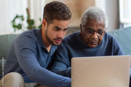 Portrait of aged man and grandson enjoying time together. African American man watching when grandson showing him pictures in computer. Taking care of senior people and modern technologies concept