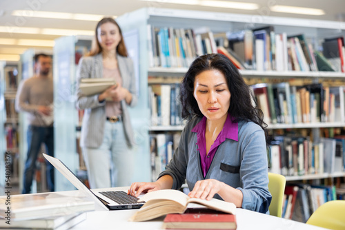 Young focused woman working with laptop and books, finding information at library and making notes