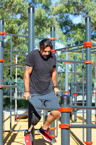 Portrait of man with artificial leg practicing on bars in summer. Handsome man training his muscles and doing push-ups on bars on sports ground. Summer sport activity of people with disability concept