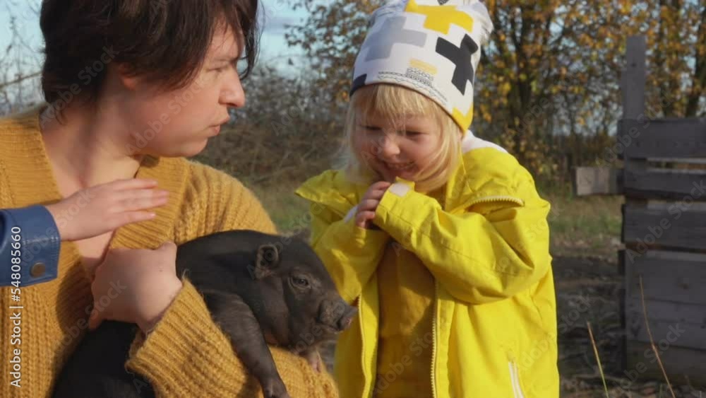 Little girl in a yellow raincoat is petting a black piglet held by a woman Little girl in a yellow raincoat is petting a black piglet held by a woman