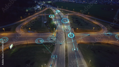 Self-driving autonomous cars move along a suburban traffic intersection in the evening. Neon HUD elements visualize the interaction of driverless cars connected to a common network photo