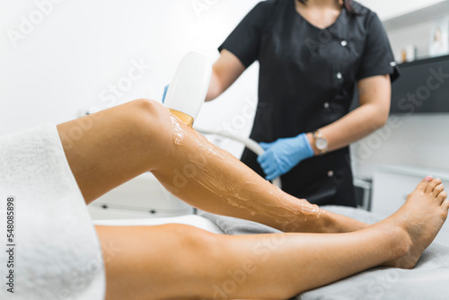 Modern ways of removing body hair. Laser hair removal. Indoor closeup shot showing legs of a Caucasian person lying down on SPA bed. One leg is covered in specialistic gel. Blurred specialist in black © PoppyPix