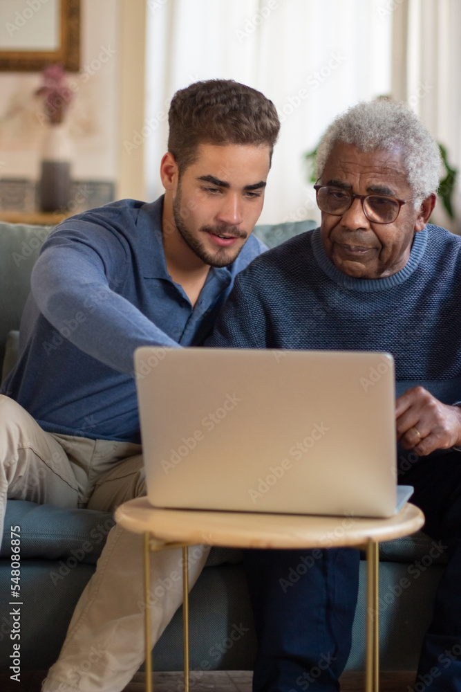 Portrait of happy granddad and his grandson resting together. Young man and granddad sitting close on sofa and watching pictures in computer. Care of senior people and modern technologies concept Portrait of happy granddad and his grandson resting together. Young man and granddad sitting close on sofa and watching pictures in computer. Care of senior people and modern technologies concept