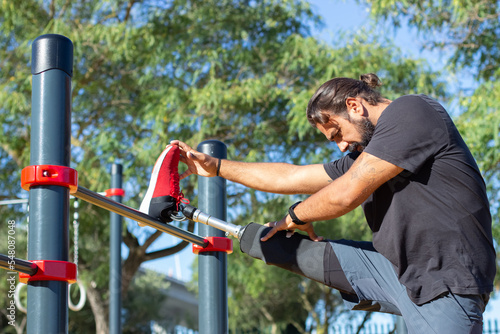 Side view of man training legs on horizontal bar outdoors. Serious man training on ground holding his prosthetic leg on horizontal bar for toning up. Sport activities of people with disability concept