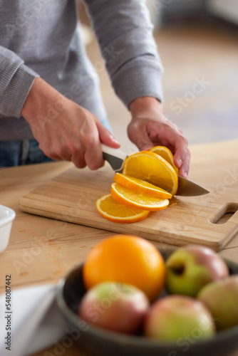 Close-up of male hands slicing big juicy orange. Unrecognizable man standing in kitchen holding orange on cutting board and preparing healthy salad for dinner. Cooking, healthy food at home concept