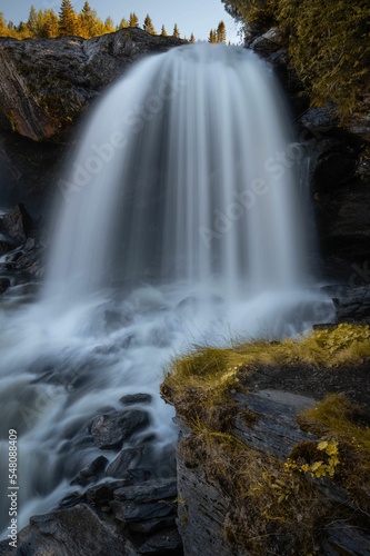 Vertical long exposure shot of Ristafallet waterfall dropping on rocks with some fall plants around photo