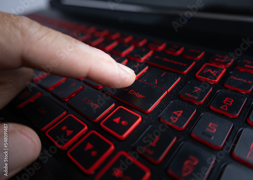 Male hand about to press the enter key on a laptop keyboard, with red lighting. (selective focus of the subject or point of action intentionally)