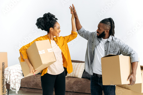 Joyful happy married african american family couple, holding boxes with things for the house, stand in living room, give five to each other, smile, rejoicing in buying their own apartment or house