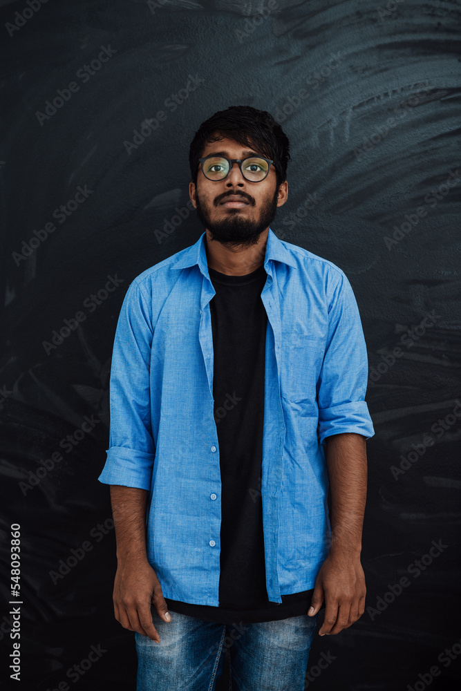 Obraz premium Indian smiling young student in blue shirt and glasses posing on school blackboard background