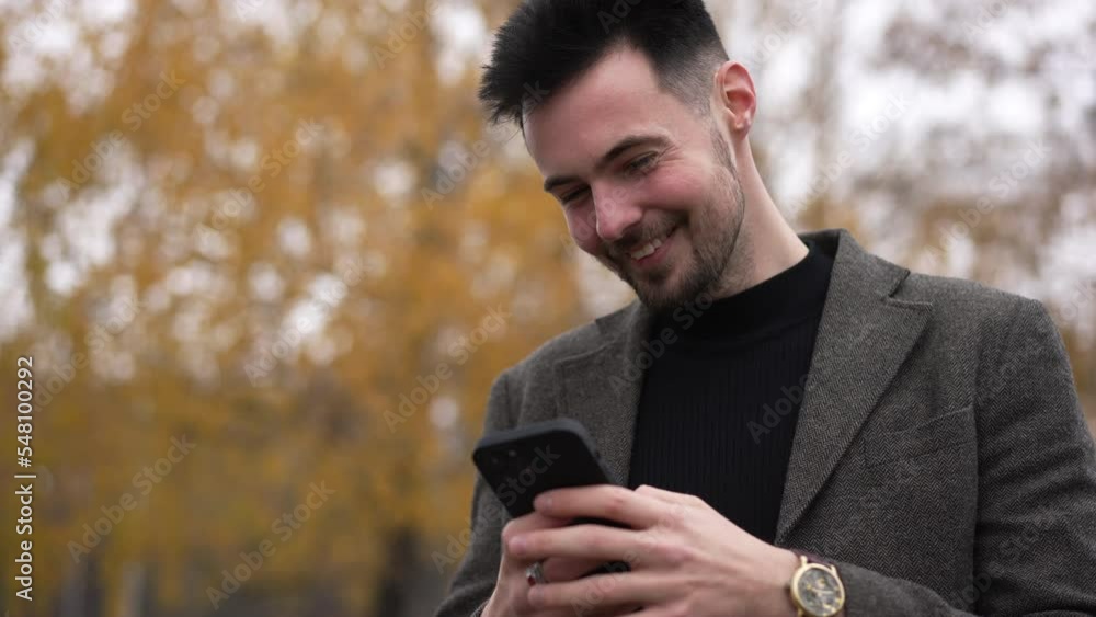 Young happy man laughing out loud messaging online in smartphone app standing on the right at background of yellow tree crown. Portrait of satisfied carefree Caucasian guy resting in forest outdoors Young happy man laughing out loud messaging online in smartphone app standing on the right at background of yellow tree crown. Portrait of satisfied carefree Caucasian guy resting in forest outdoors