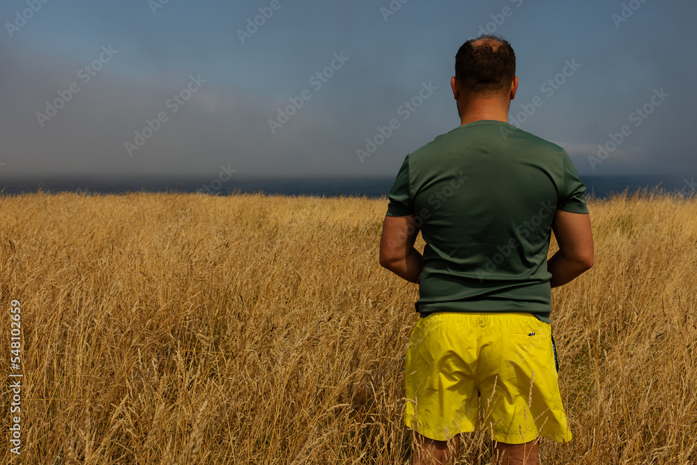 A man stands with his back in a field of yellow grass staring at the blue ocean in the distance. Green jersey and yellow bright shorts.