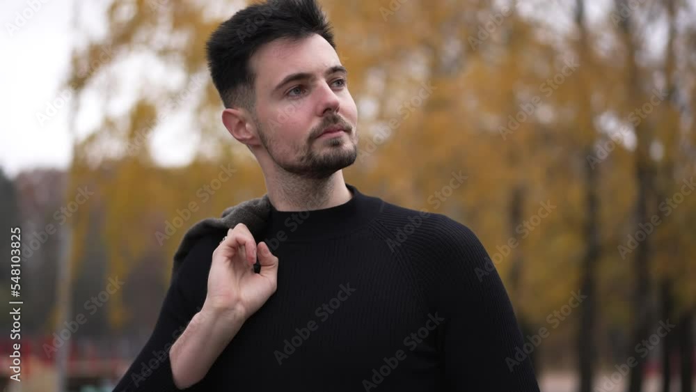 Confident young guy with blazer on shoulder standing in autumn park checking time on watch. Portrait of Caucasian handsome man looking at camera smiling waiting outdoors Confident young guy with blazer on shoulder standing in autumn park checking time on watch. Portrait of Caucasian handsome man looking at camera smiling waiting outdoors