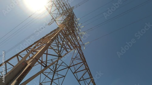 Looking up along large electrical tower and power lines overhead against sky and bright sun lens flare, tilt up, rotate; in Ontario, Canada.