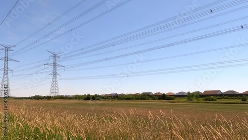 Large electrical towers and power lines over empty field with houses in distance, pan right; in Ontario Canada.