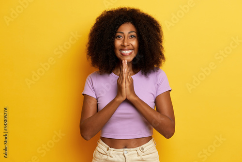 Positive charming curly haired woman holds palms pressed together and smiles widely, poses over yellow background photo