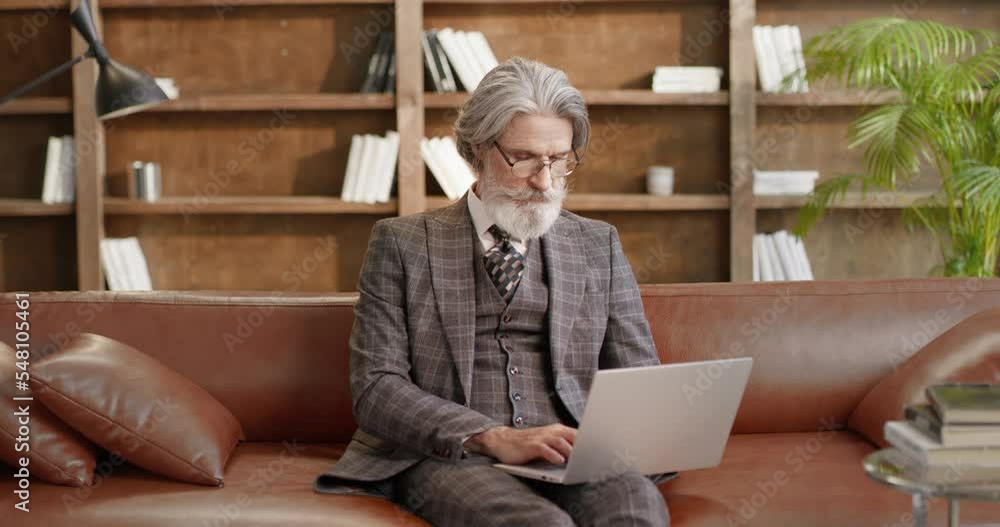 Mature focused serious man working online on laptop typing wearing suit. Grey-haired elderly stylish male sitting on sofa wearing glasses using computer technologies. Working online concept. Mature focused serious man working online on laptop typing wearing suit. Grey-haired elderly stylish male sitting on sofa wearing glasses using computer technologies. Working online concept.