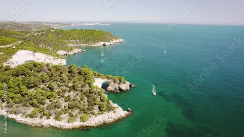 Drone flying upwards and in circulair motion over the coastline of Gargano national park showing an arch in the sea and boats passing by in 4k photo